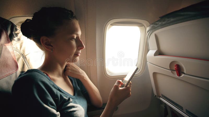 Donna turistica che si siede vicino alla finestra dell'aeroplano al tramonto e che per mezzo del telefono cellulare durante il vo immagine stock libera da diritti Donna turistica che si siede vicino alla finestra dell'aeroplano al tramonto e che per mezzo del telefono cellulare durante il vo immagine stock libera da diritti
