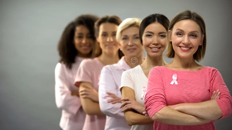 Donne sorridenti in camice rosa con i nastri del cancro al seno che stanno nella fila, supporto
