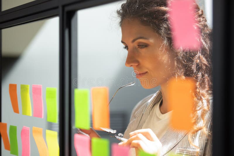 Head shot close up thoughtful young female boss team leader looking at kanban scrum glass window board, developing start up project marketing strategy, managing workflow, checking tasks at office. Head shot close up thoughtful young female boss team leader looking at kanban scrum glass window board, developing start up project marketing strategy, managing workflow, checking tasks at office.