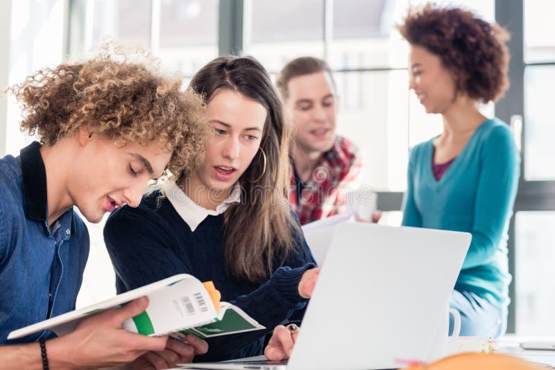 Dos estudiantes jovenes que buscan para una respuesta a su pregunta en línea y en un libro de texto mientras que se sienta en el escritorio en la sala de clase durante rotura