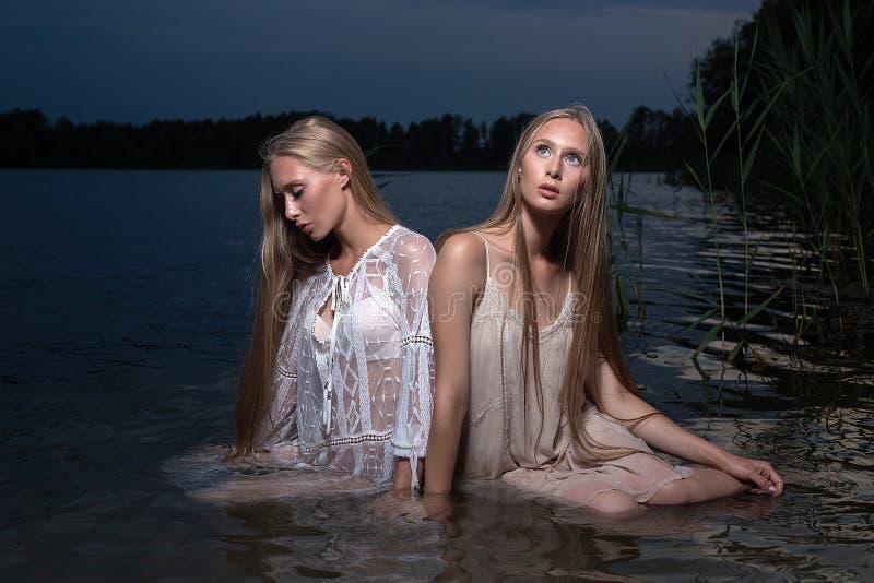 Dos hermanas gemelas jóvenes posando con vestidos de luz en el agua del lago en la noche de verano foto de archivo