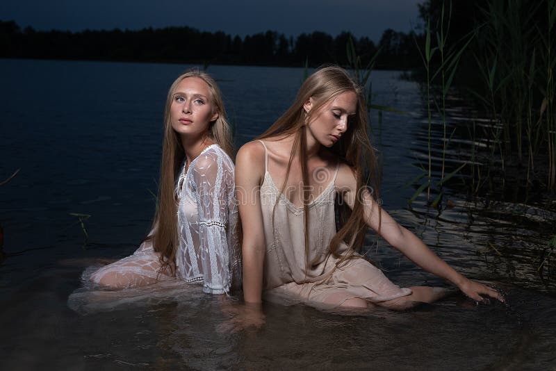 Dos hermanas gemelas jóvenes posando con vestidos de luz en el agua del lago en la noche de verano fotografía de archivo libre de regalías