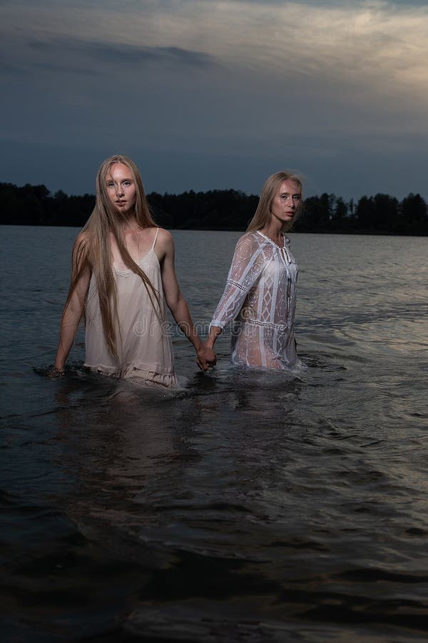 Dos hermanas gemelas jóvenes posando con vestidos de luz en el agua del lago en la noche de verano foto de archivo libre de regalías