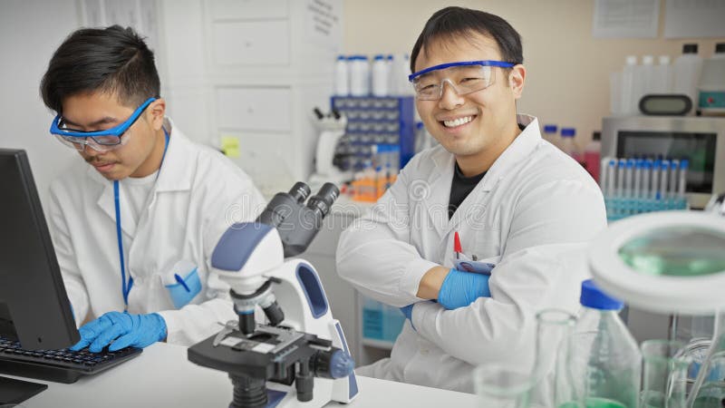 Dos hombres asiáticos con abrigos blancos trabajando en un laboratorio de ciencias uno usando una computadora y el otro sonriendo fotografía de archivo libre de regalías