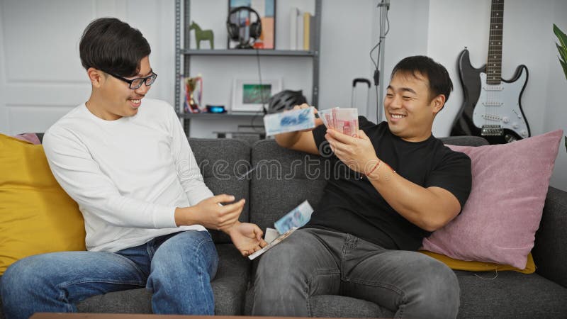 Dos hombres asiáticos ríen al comparar los billetes de yuan en una acogedora sala de estar con una guitarra al fondo foto de archivo