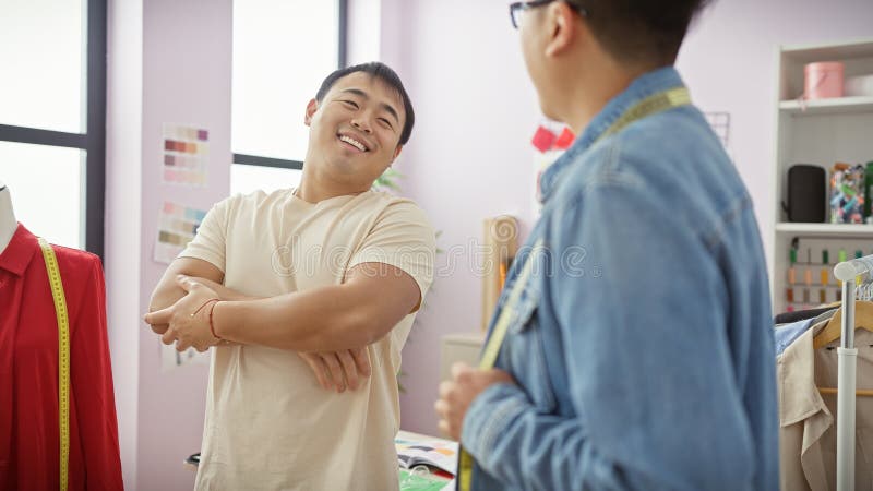 Dos hombres asiáticos trabajan juntos en una tienda de sastre brillante discutiendo prendas con una cooperación sonriente imagenes de archivo
