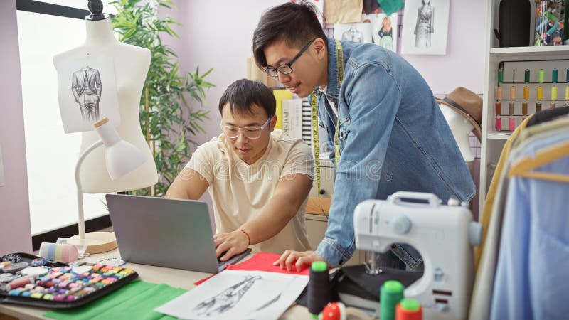 Dos hombres colaboran en una tienda de sastre examinando los diseños de una laptop entre telas y equipos de costura foto de archivo libre de regalías