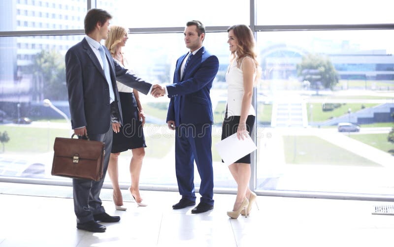 Dos hombres de negocios confiados que sacuden las manos y que sonríen mientras que se coloca en la oficina así como el grupo de c foto de archivo libre de regalías Dos hombres de negocios confiados que sacuden las manos y que sonríen mientras que se coloca en la oficina así como el grupo de c foto de archivo libre de regalías