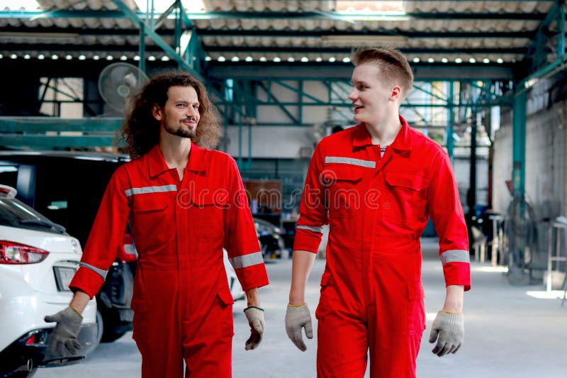 Dos hombres mecánicos guapos con uniforme rojo que caminan por fila de vehículo en el taller mecánico del garaje amigo trabajan ju foto de archivo