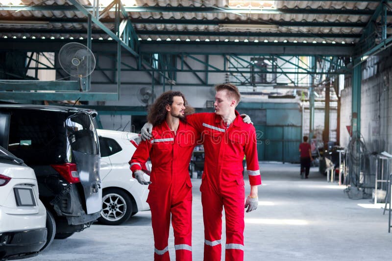 Dos hombres mecánicos guapos con uniforme rojo que caminan por fila de vehículo en el taller mecánico del garaje amigo trabajan ju imagen de archivo