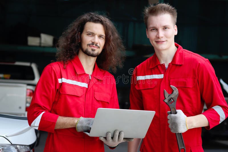 Dos hombres mecánicos guapos con uniforme rojo sujetando una computadora portátil durante la discusión en conjunto en un técnico d fotografía de archivo libre de regalías