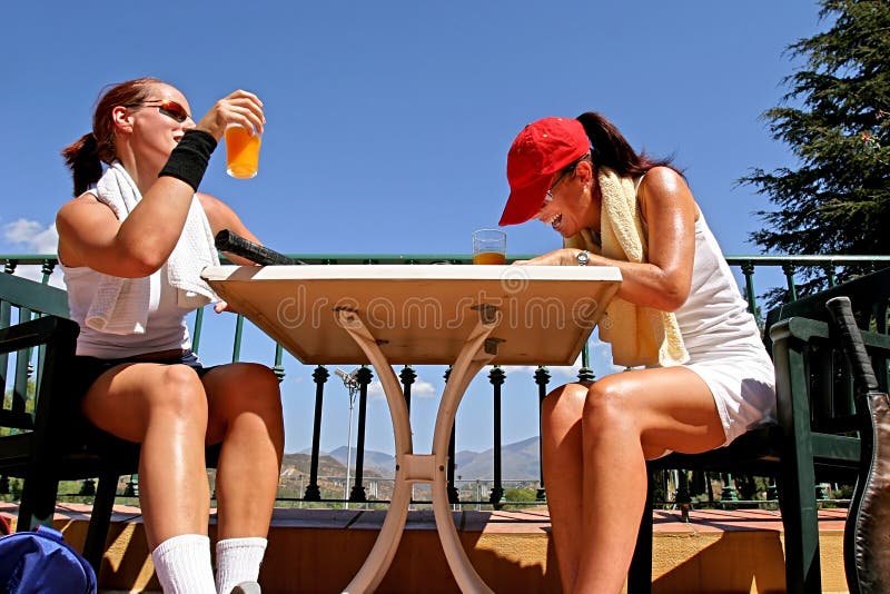 Dos jugadores de tenis de sexo femenino que comparten una broma después de un juego. Goce de un vidrio de zumo de naranja en el so fotos de archivo libres de regalías Dos jugadores de tenis de sexo femenino que comparten una broma después de un juego. Goce de un vidrio de zumo de naranja en el so fotos de archivo libres de regalías