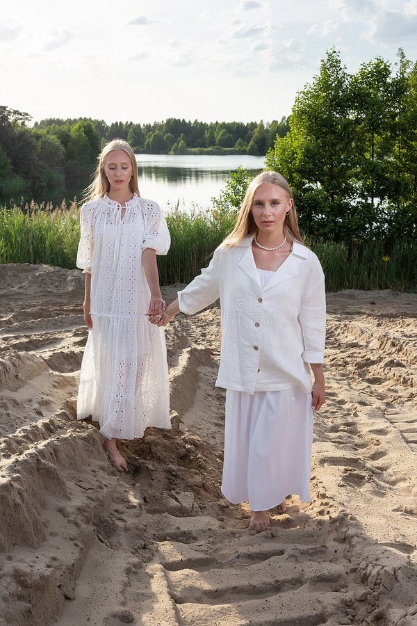 Dos jóvenes hermanas gemelas atractivas posando en la playa de arena con elegantes ropas blancas fotos de archivo