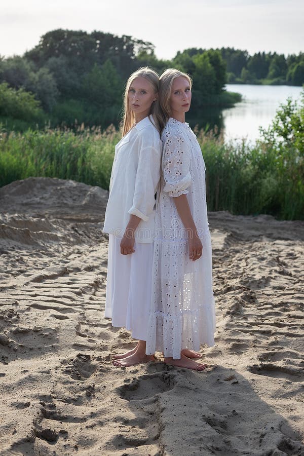 Dos jóvenes hermanas gemelas atractivas posando en la playa de arena con elegantes ropas blancas imagenes de archivo