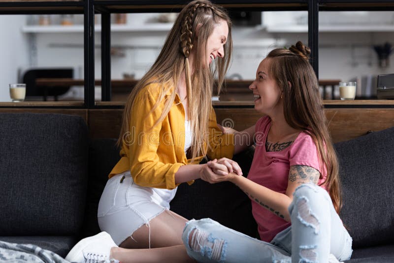 Dos Lesbianas Sonrientes Cogidas De La Mano Mientras Se Sientan En El Sofá De La Sala De Estar