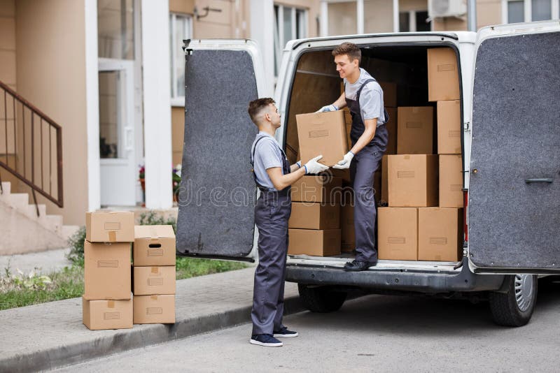 Dos motores hermosos jovenes que llevan los uniformes están descargando la furgoneta por completo de cajas Movimiento de la casa, imagen de archivo Dos motores hermosos jovenes que llevan los uniformes están descargando la furgoneta por completo de cajas Movimiento de la casa, imagen de archivo
