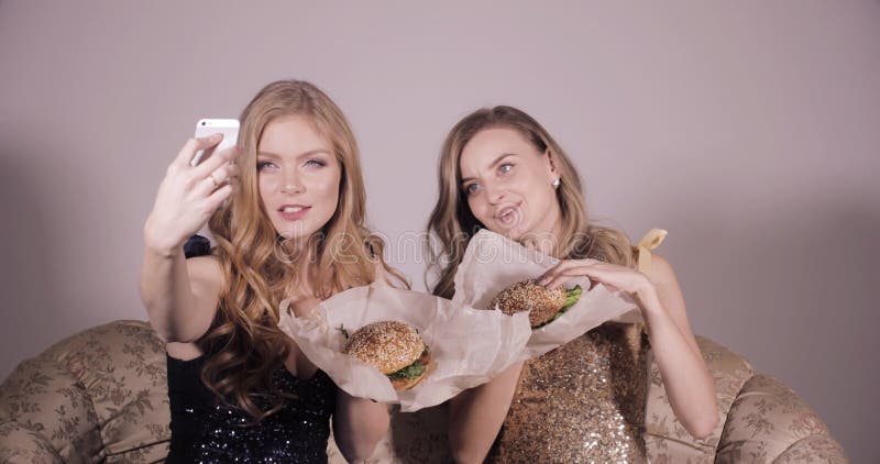 Dos muchachas hermosas que sostienen las hamburguesas y que hacen el selfie