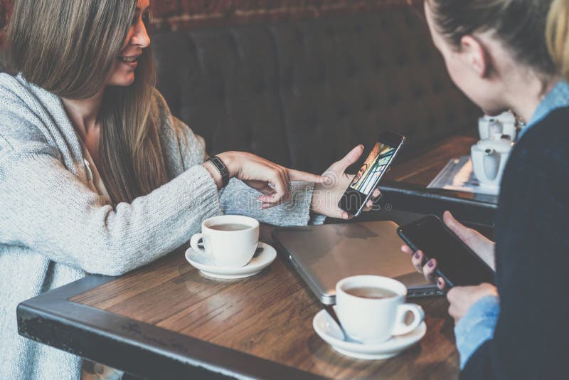 Dos mujeres de negocios jovenes que se sientan en la tabla y que usan smartphones Mujer que muestra imagen del colega en la panta fotos de archivo Dos mujeres de negocios jovenes que se sientan en la tabla y que usan smartphones Mujer que muestra imagen del colega en la panta fotos de archivo