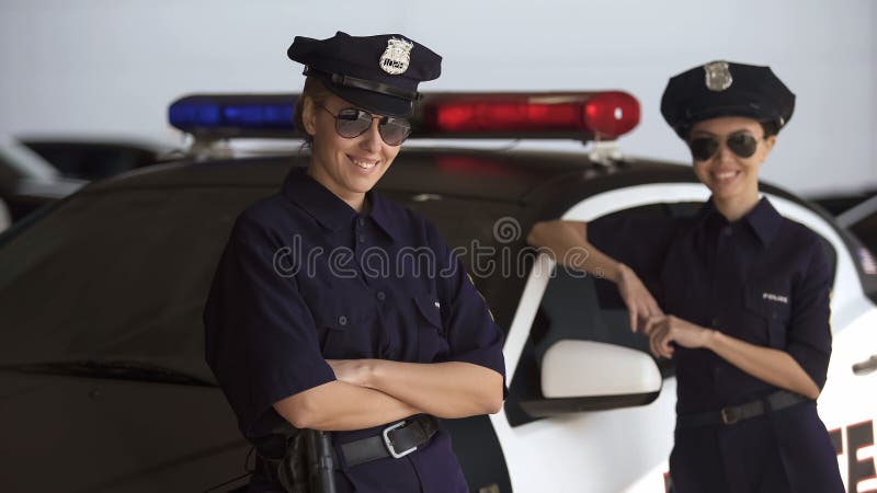 Dos mujeres en los uniformes de la policía que se colocan cerca del coche patrulla y que sonríen, ley y orden imagenes de archivo Dos mujeres en los uniformes de la policía que se colocan cerca del coche patrulla y que sonríen, ley y orden imagenes de archivo