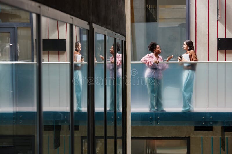 Dos mujeres sonrientes charlando de pie en el segundo piso del edificio de oficinas. Toma en gran angular con composición geométrica de dos mujeres sonrientes participando en una conversación mientras están en el segundo piso del espacio de copia del edificio de oficinas contemporáneo