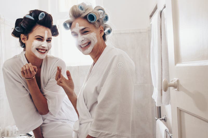 Dos mujeres sonrientes en las albornoces que aplican la crema de cara imagen de archivo Dos mujeres sonrientes en las albornoces que aplican la crema de cara imagen de archivo