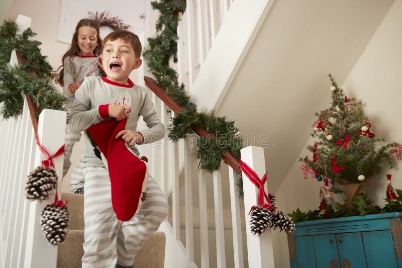 Dos niños emocionados que llevan los pijamas que corren abajo de las escaleras que sostienen medias el mañana de la Navidad foto de archivo Dos niños emocionados que llevan los pijamas que corren abajo de las escaleras que sostienen medias el mañana de la Navidad foto de archivo