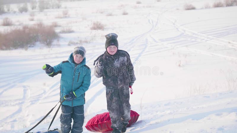Dos niños montan en una colina nevosa en un trineo Los niños suben al top de la montaña Paseos en el aire fresco