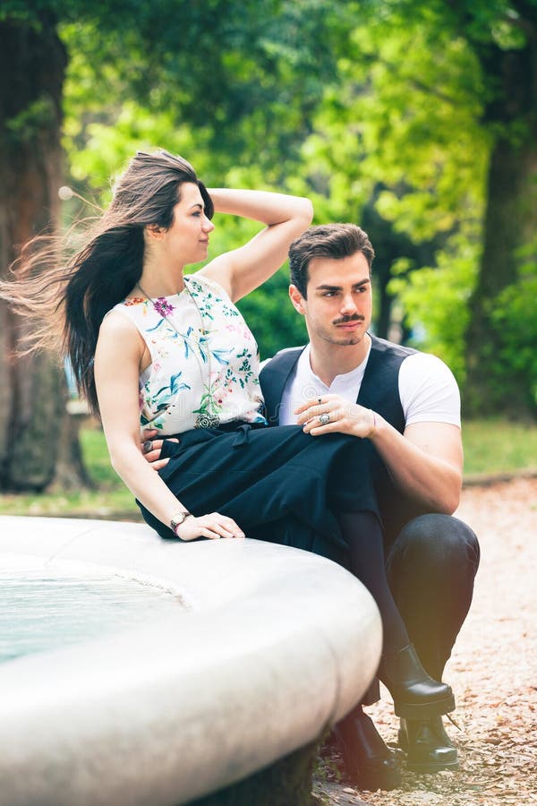 A beautiful young couple is relaxing in a park with trees and vegetation. The girl is sitting on a fountain. The young men crouched in front of her. Hair in the wind. A beautiful young couple is relaxing in a park with trees and vegetation. The girl is sitting on a fountain. The young men crouched in front of her. Hair in the wind.