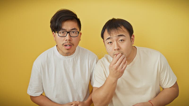 Dos sorprendidos hombres asiáticos parados juntos contra un vibrante fondo amarillo que invoca curiosidad y asombro imagen de archivo