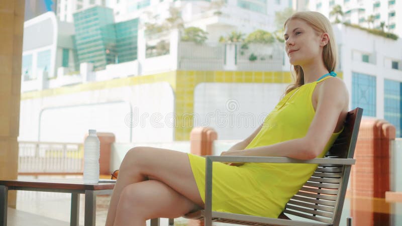 Dosyć marzycielska młoda kobieta w kolor żółty sukni obsiadaniu w krześle blisko pływackiego basenu w mieście