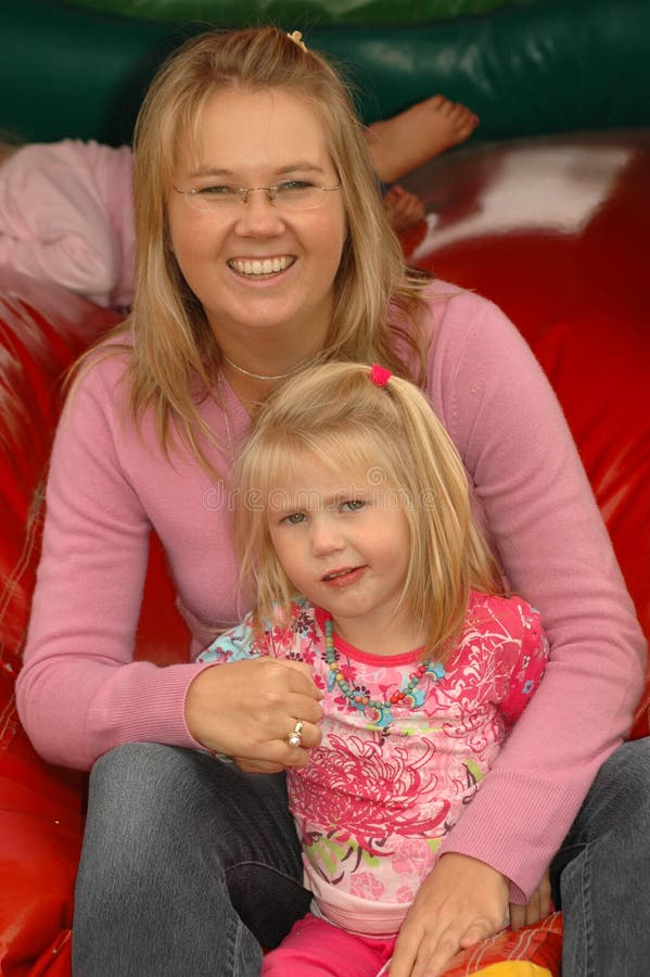 A portrait of a young caucasian mother together with her blond little grogeous white daughter with cute expression in the face sitting on a jumping castle, smiling, having great fun and enjoying a birthday party outdoors. A portrait of a young caucasian mother together with her blond little grogeous white daughter with cute expression in the face sitting on a jumping castle, smiling, having great fun and enjoying a birthday party outdoors