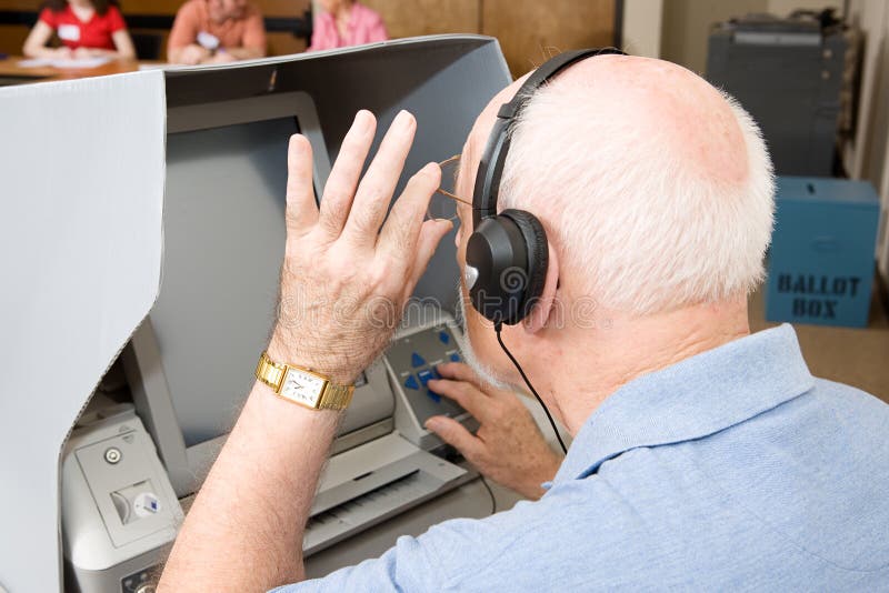 Senior man using a new touch screen voting machine equipped for hearing and vision impaired. Election workers visible in background. Senior man using a new touch screen voting machine equipped for hearing and vision impaired. Election workers visible in background.