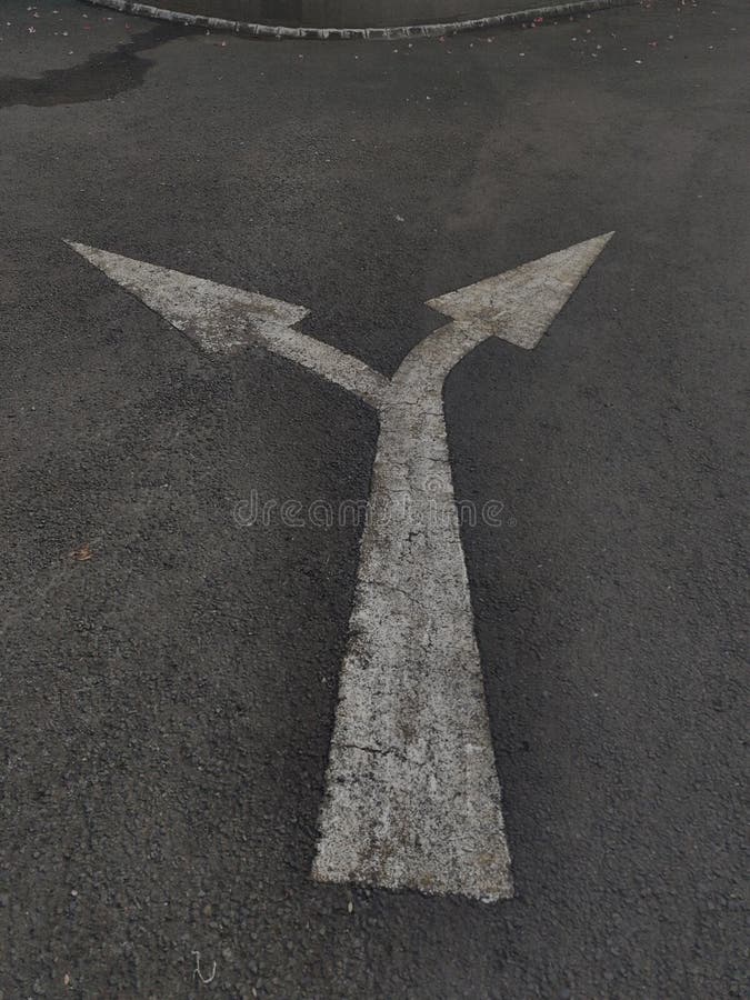 White double-headed directional arrow painted on dark asphalt. The arrow splits into two directions, indicating a choice between left and right. The road surface shows signs of wear and age. The surrounding area includes a slight curve in the road boundary marked by concrete edging. The setting appears to be an urban or suburban street environment. Painted curve stock images, royalty-free photos and pictures