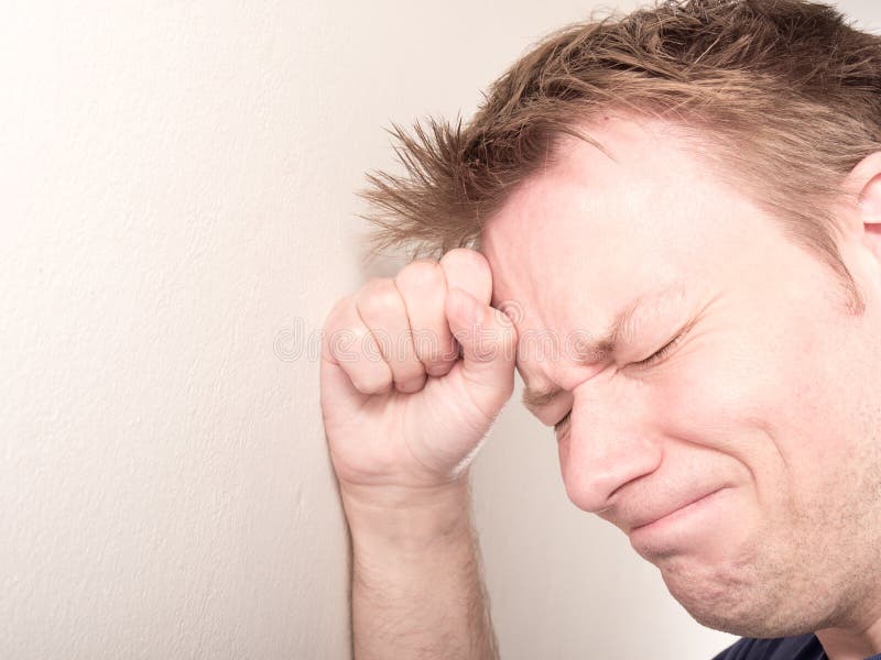 Young sad man leaning his head against his clenched fist on a white wall with copy space to the left - for your suffering, depression, grief and death concepts. Young sad man leaning his head against his clenched fist on a white wall with copy space to the left - for your suffering, depression, grief and death concepts