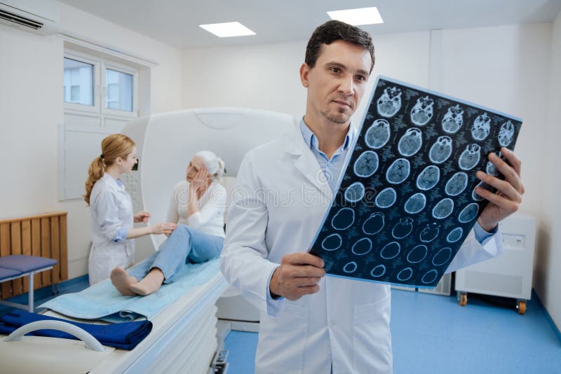 Photographs of human brain. Confident pleasant male doctor working with MRI scan results and putting a diagnosis while doing his job. Photographs of human brain. Confident pleasant male doctor working with MRI scan results and putting a diagnosis while doing his job