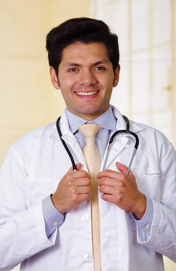 Doutor de sorriso considerável seguro que levanta e que olha a câmera com um estetoscópio em torno de seu pescoço, no fundo do es imagem de stock Doutor de sorriso considerável seguro que levanta e que olha a câmera com um estetoscópio em torno de seu pescoço, no fundo do es imagem de stock
