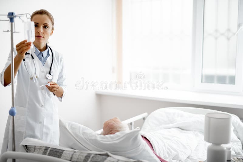 Doutor novo que ajusta a garrafa da infusão ao estar perto da cama paciente foto de stock Doutor novo que ajusta a garrafa da infusão ao estar perto da cama paciente foto de stock