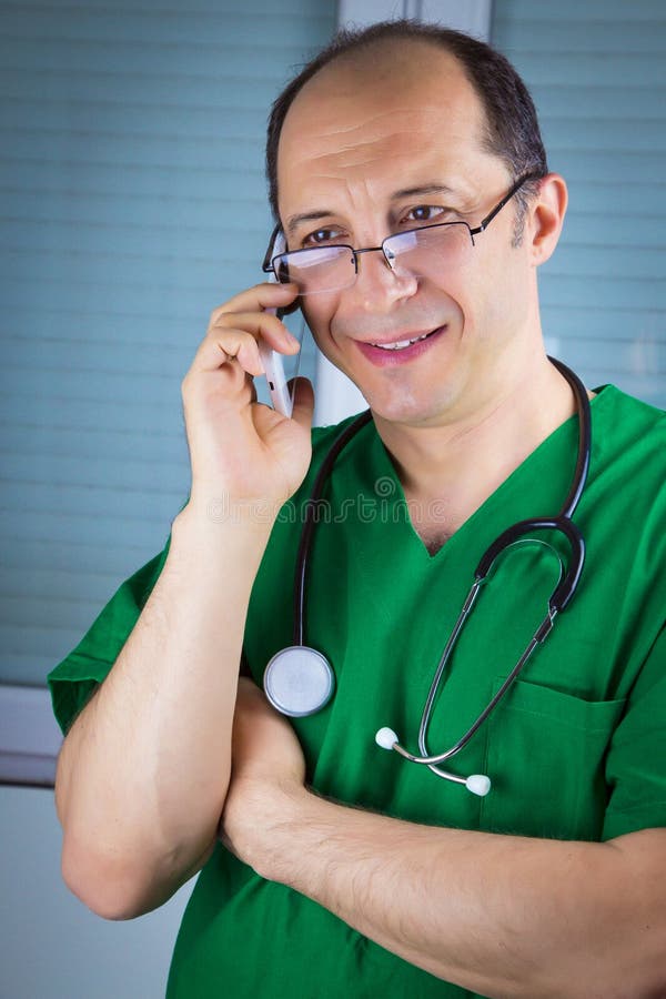 Doctor talking on cell phone. Stetoscope on his nack. Wearing green uniform and glasses. Doctor talking on cell phone. Stetoscope on his nack. Wearing green uniform and glasses