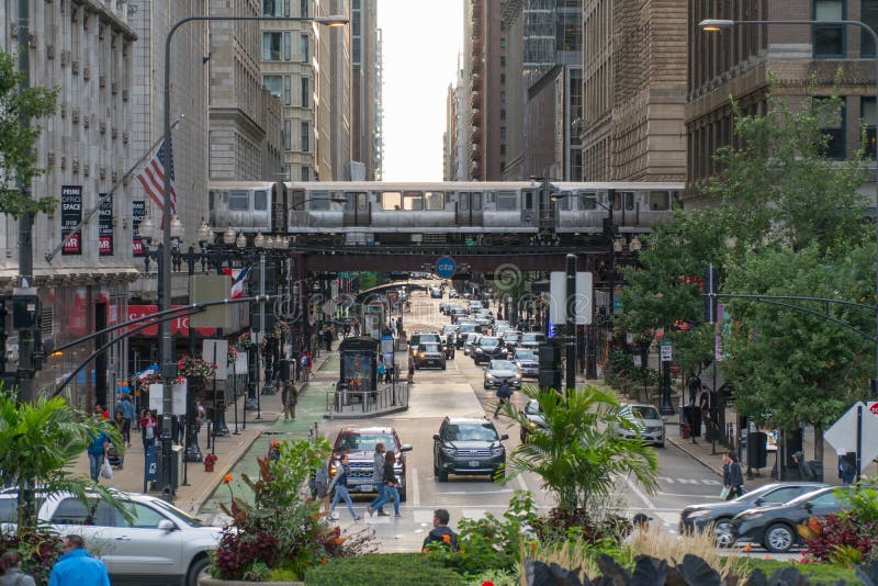 Chicago, IL, USA - Circa 2019: View down busy street in downtown as Loop train passes overhead tracks over avenue morning rush hour traffic and crowds of people walking down sidewalks to work. Chicago, IL, USA - Circa 2019: View down busy street in downtown as Loop train passes overhead tracks over avenue morning rush hour traffic and crowds of people walking down sidewalks to work