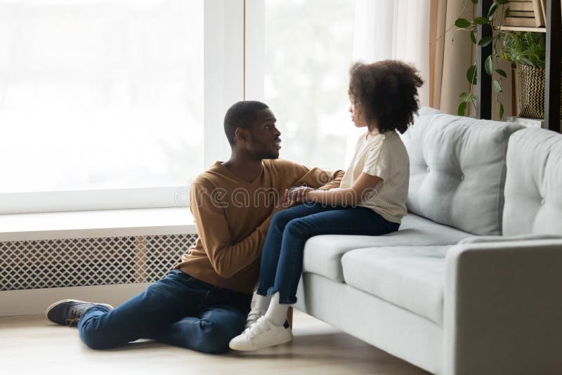 Caring African American father comforting, supporting upset adorable preschool daughter, sitting on floor, holding hands, loving dad talking to stressed child girl, empathy and protection concept. Caring African American father comforting, supporting upset adorable preschool daughter, sitting on floor, holding hands, loving dad talking to stressed child girl, empathy and protection concept