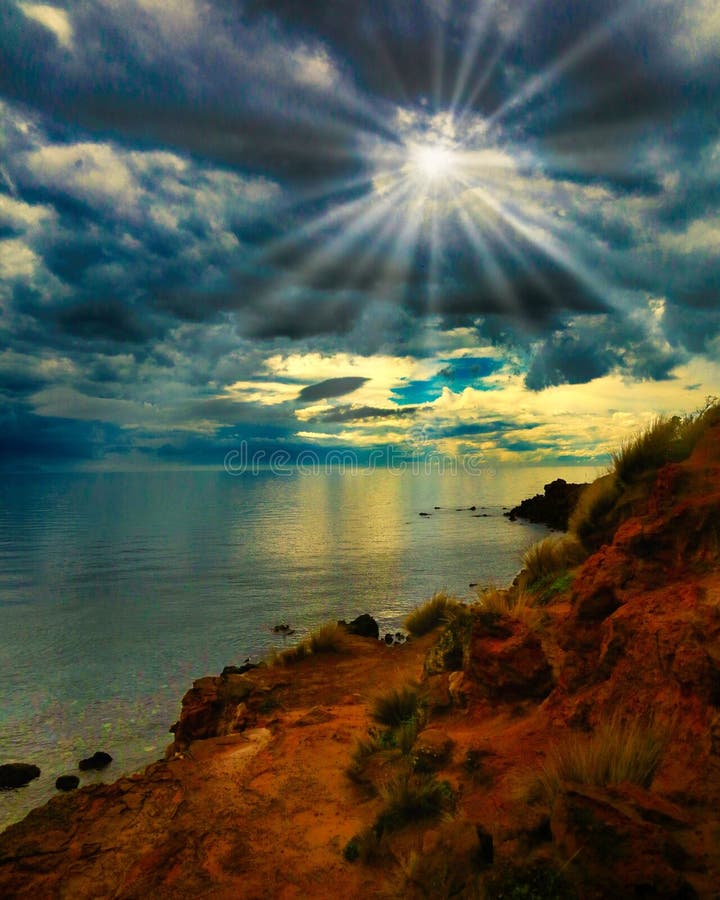 Coastal landscape with sun rays piercing through clouds over a calm sea, rocky shore in foreground stock photos