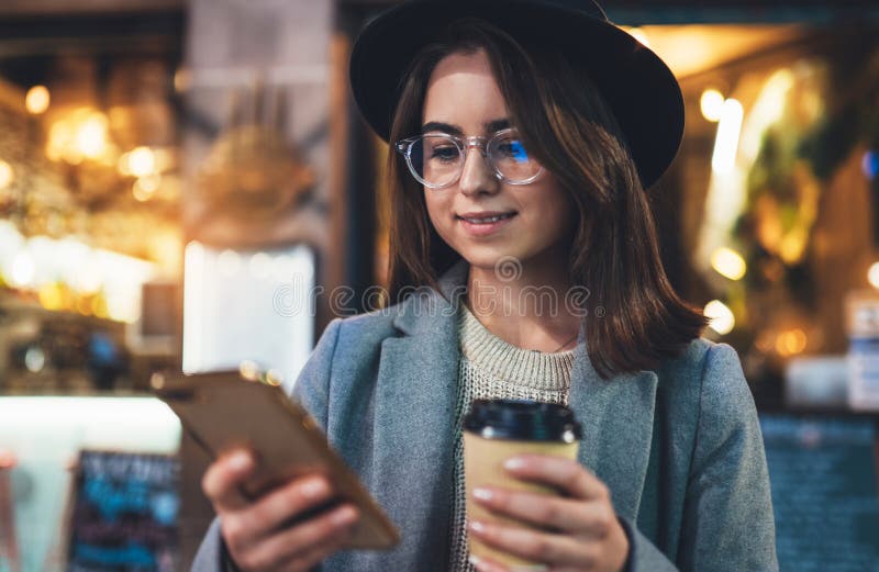 Draußen Lächelnmädchen-Getränktasse kaffee mithilfe des mobilen Smartphone auf hellem Hintergrund bokeh Nachtstadt-Frauengriff im lizenzfreies stockfoto