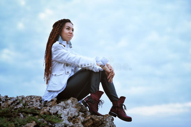Dreadlocks fashionable girl posing near sea at sunset, sits on high stone on sky background royalty free stock photography Dreadlocks fashionable girl posing near sea at sunset, sits on high stone on sky background royalty free stock photography