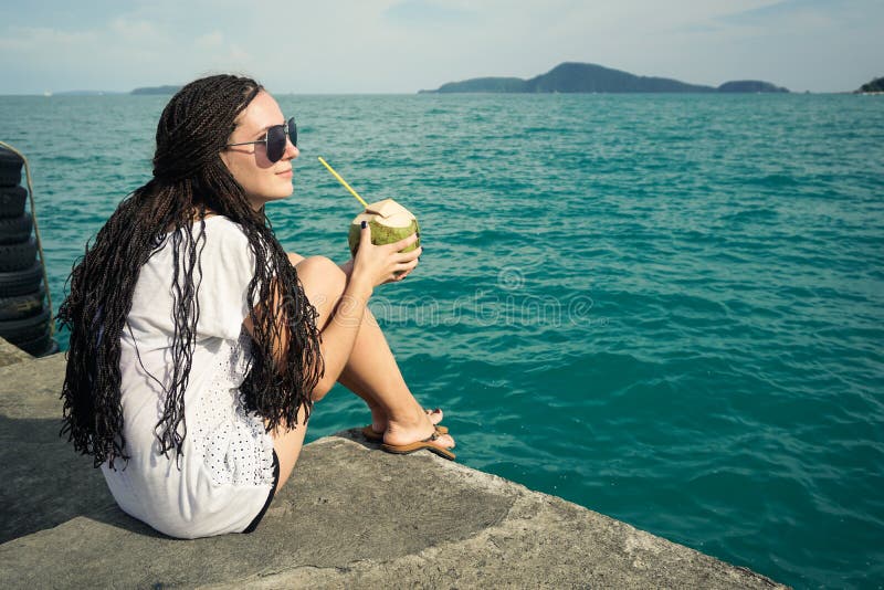 Posteriore da giovane donna a lungo trecce possesso noce di cocco paglia un cercando lontananza Oceano mentre sul molo.