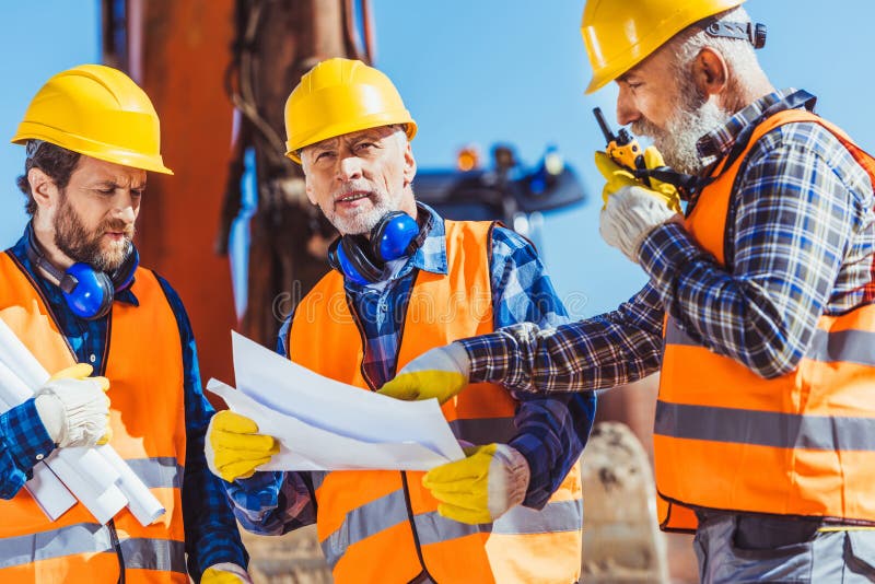 Drei Arbeitskräfte, die Baupläne prüfen und auf portablem Radio sprechen stockfotografie Drei Arbeitskräfte, die Baupläne prüfen und auf portablem Radio sprechen stockfotografie