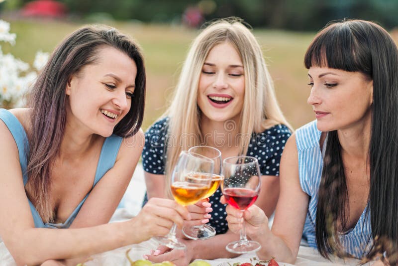 Drei junge Frauen in den blauen Kleidern und in den Hüten liegen auf Plaid und Getränkwein Picknick im Freien auf Gras auf Strand stockfoto