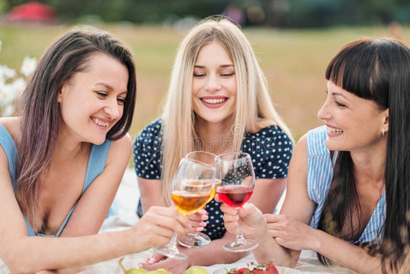 Drei junge Frauen in den blauen Kleidern und in den Hüten liegen auf Plaid und Getränkwein Picknick im Freien auf Gras auf Strand lizenzfreie stockfotos