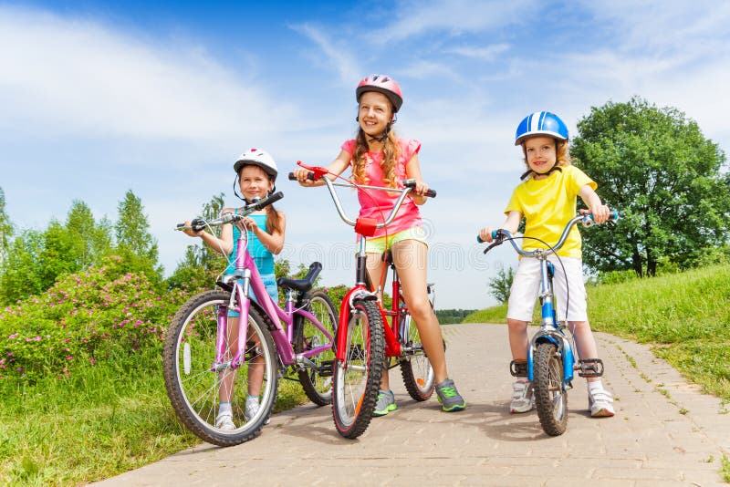 Three girls on a paved road stand holding with bicycles on sunny summer day . Three girls on a paved road stand holding with bicycles on sunny summer day .