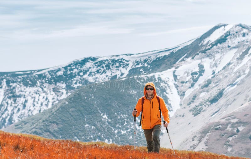 Oblečená jasne oranžová bunda batohom kráčajúca po červenej čučoriedkovej oblasti pomocou trekingových palíc s pozadím pohoria,