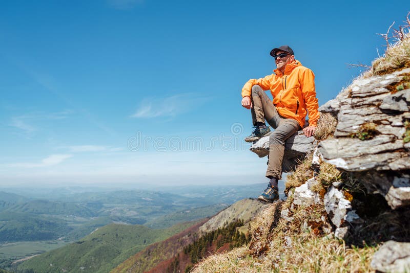 Oblečený turista v žiarivo oranžovej bunde v baseballovej čiapke a slnečných okuliaroch sedí na skalnom útese a užíva si zelené údolie pri malej fatre